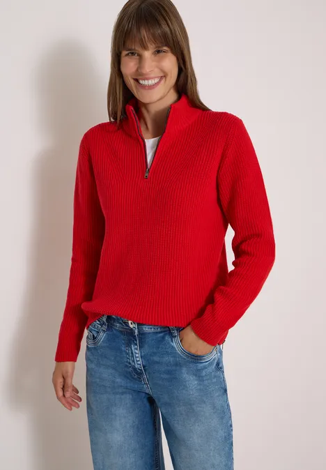 Eine lächelnde Frau trägt einen roten Pullover und blaue Jeans vor weißem Hintergrund.