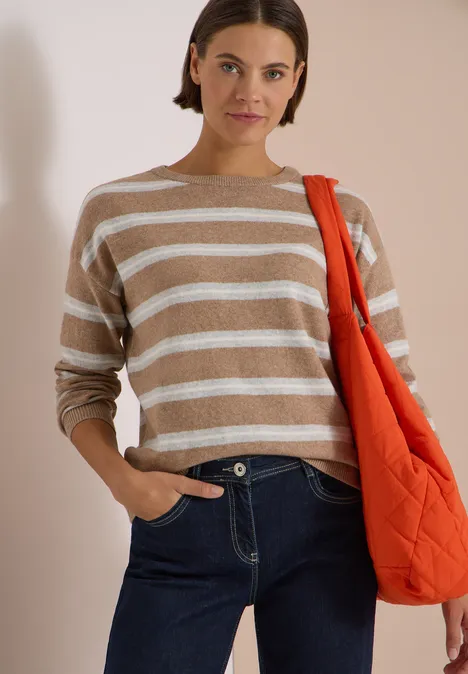 Frau in beige-weiß gestreiftem Pullover, Jeans und oranger Umhängetasche vor hellem Hintergrund.