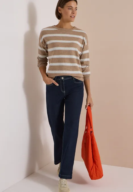 Eine Frau steht vor neutralem Hintergrund, trägt gestreiften Pullover, weite Jeans und hält eine orangefarbene Tasche.