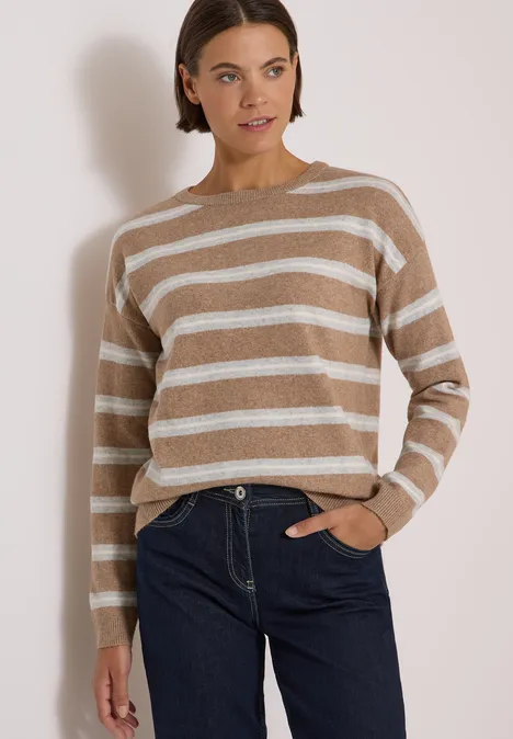 Frau in beige-weißgestreiftem Pullover und blauer Jeans, eine Hand in der Tasche, vor heller Wand.