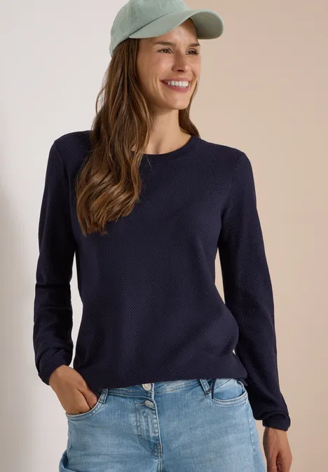 Eine lächelnde Frau trägt einen dunkelblauen Pullover, Jeans und eine hellgrüne Baseballmütze.