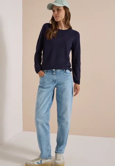 Frauenmodell in dunkelblauem Pullover, hellblauen Jeans und Mütze steht vor neutralem Hintergrund.