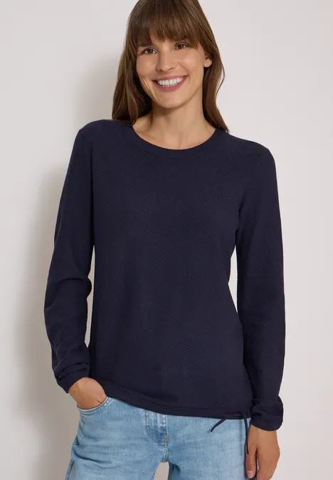 Frau lacht in dunkelblauem Pullover und Jeans vor weißem Hintergrund.
