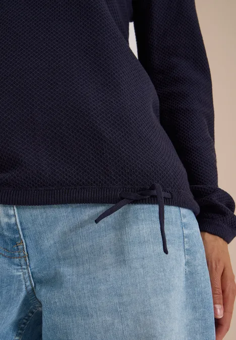 Dunkelblauer, texturierter Pullover mit seitlicher Kordel am Saum, kombiniert mit hellblauen Jeans.