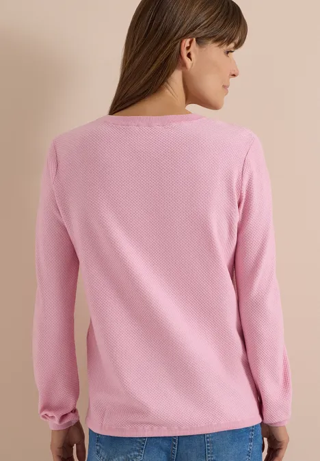 Rückansicht einer Frau mit rosa Langarmpullover und blauer Jeans vor neutralem Hintergrund.