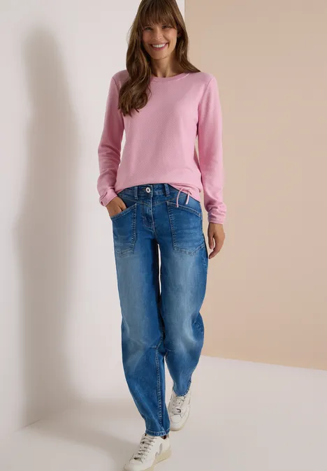 Frau im rosafarbenen Pullover, blauen Jeans und weißen Sneakers vor neutralem Hintergrund.