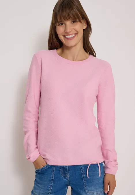 Eine lächelnde Frau in rosa Langarmshirt und Jeans, Hände in den Taschen vor neutralem Hintergrund.