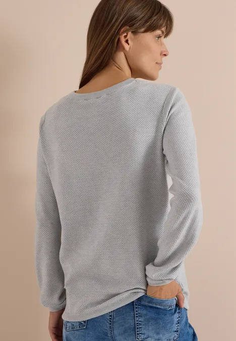 Frau von hinten in grauem Langarmshirt und Jeans vor beigem Hintergrund.