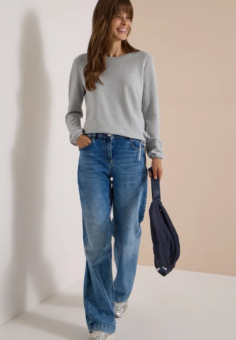 Frau in grauem Langarmshirt, weiten Jeans und Sneakers trägt eine dunkelblaue Tasche und lächelt.
