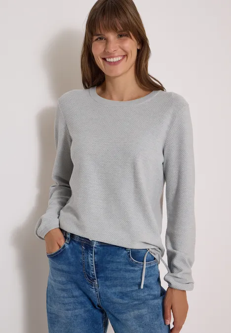 Frau in grauem Pullover und Jeans, lächelnd vor hellem Hintergrund mit einer Hand in der Tasche.
