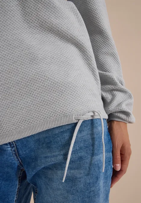 Nahaufnahme eines grauen Waffelstrick-Pullis mit Kordelzug am Saum, kombiniert mit blauen Jeans.