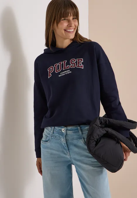 Frau trägt lässigen dunkelblauen Hoodie mit „PULSE“-Schriftzug, Jeanshose und hält eine schwarze Steppjacke.