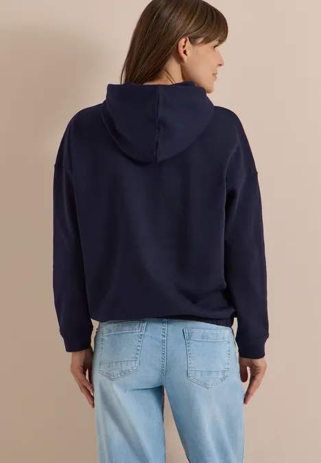 Rückenansicht einer Frau in dunkelblauer Kapuzenjacke und hellblauen Jeans.