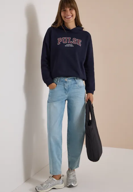Eine Frau trägt ein dunkelblaues Sweatshirt mit 'PULSE', hellblaue Jeans, graue Sneakers und eine schwarze Tasche.