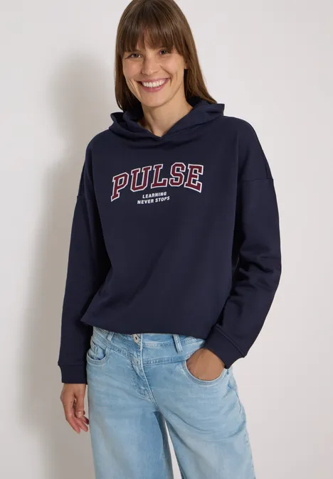Frau in dunkelblauer Kapuzenjacke mit 'PULSE'-Aufdruck und blauen Jeans, die vor hellem Hintergrund lächelt.
