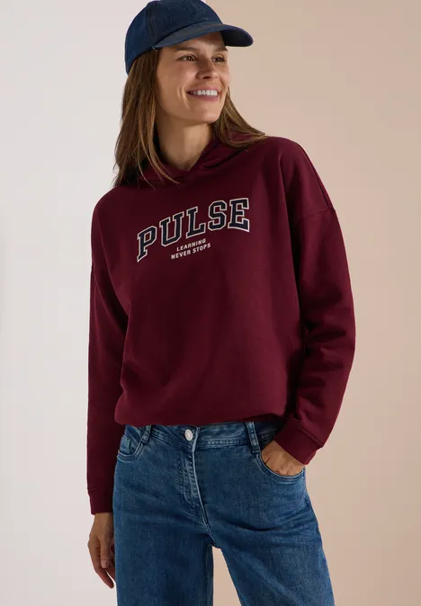 Lächelnde Frau mit burgunderroter Kapuzenjacke mit 'PULSE'-Schriftzug, Jeans und Baseballcap.