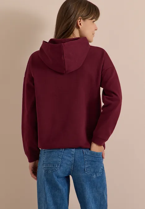 Rückansicht einer Frau in Jeans und bordeauxrotem Kapuzenpullover vor hellem Hintergrund.