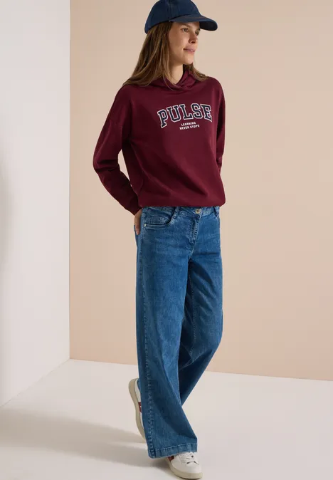 Frau trägt lässiges Outfit mit burgunderrotem Pullover, blauer Jeanshose, Sneakern und Cap.