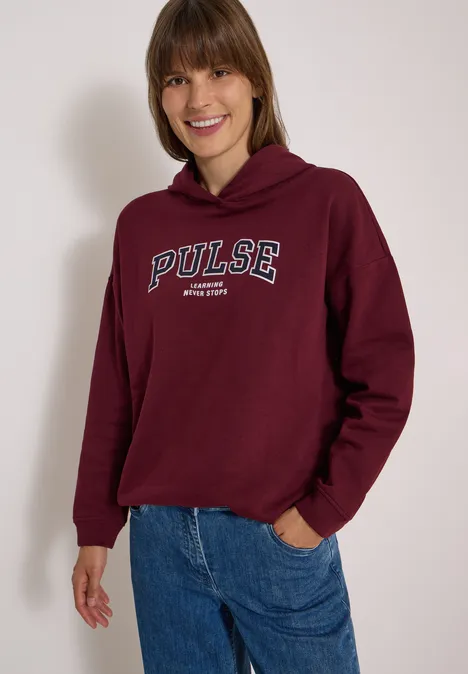 Junge Frau in weinrotem Hoodie mit 'PULSE'-Schriftzug und blauen Jeans lächelt vor hellem Hintergrund.