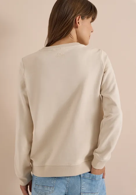 Eine Frau mit braunen Haaren trägt einen hellbeigen Pullover und blaue Jeans, Rückansicht.