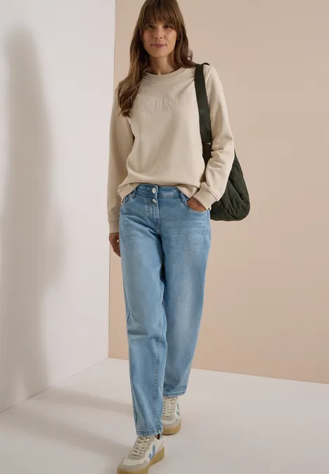 Frau mit beigem Sweater, blauen Jeans und Sneakers trägt grüne Tasche vor hellbeiger Wand.