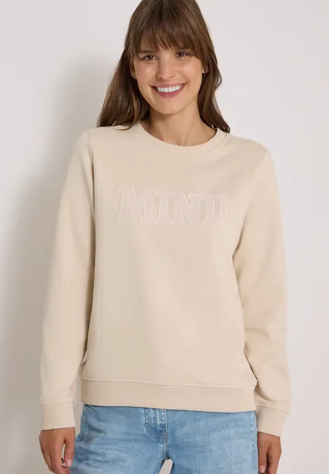 Frau in beige Sweatshirt mit Schriftzug 'MIND' und blauer Jeans, lächelnd vor hellem Hintergrund.