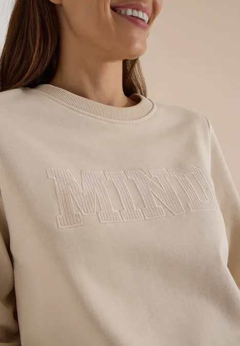 Frau im Profil trägt cremefarbenes Sweatshirt mit gesticktem Schriftzug „MIND“ und lächelt leicht.