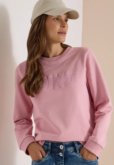 Frau trägt rosa Sweatshirt, blaue Jeans und beige Baseballkappe.