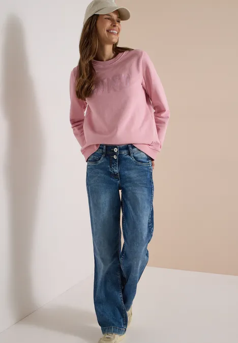 Junge Frau mit rosa Pullover, blauen Jeans und weißem Cap lehnt lächelnd an einer Wand.