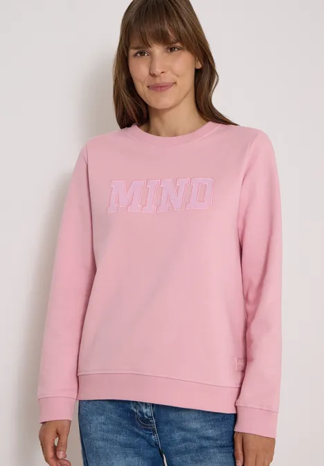 Frau in rosa Sweatshirt mit weißem "MIND"-Schriftzug, lächelt.