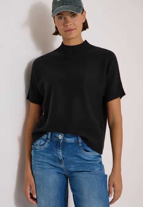 Frau in schwarzem Kurzarm-Oberteil und blauen Jeans vor weißem Hintergrund.