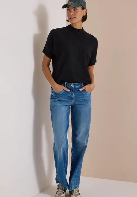 Frau in schwarzem Kurzarmshirt, blauen Jeans und Baseballkappe lehnt an heller Wand.
