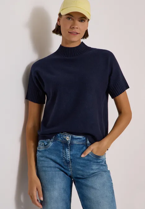 Frau trägt marineblaues Kurzarm-Strickshirt, Jeans und hellgelbe Kappe vor hellem Hintergrund.