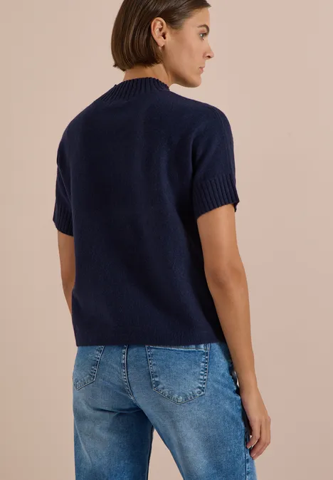 Frau von hinten in marineblauem Kurzarm-Pullover und blauen Jeans vor beigem Hintergrund.
