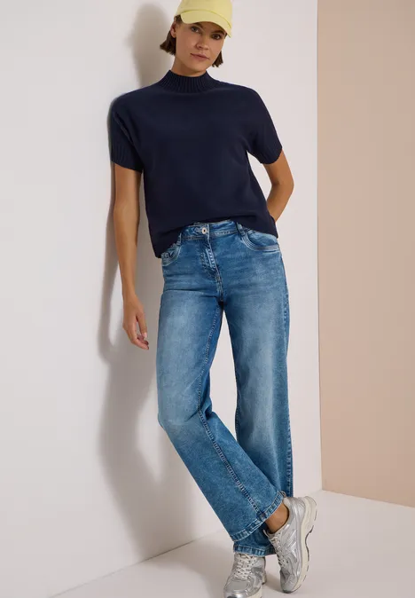 Frau lehnt entspannt an Wand, trägt navyblaues T-Shirt, gerade geschnittene Jeans, weiße Sneaker und gelbe Cap.