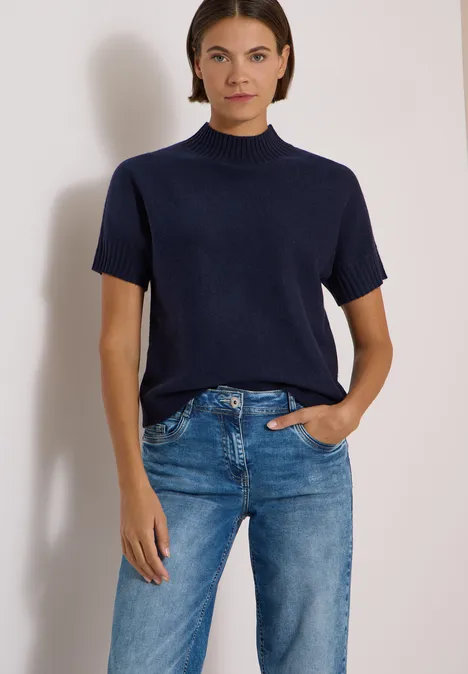 Frau in blauem kurzärmligem Rollkragenpullover und geraden blauen Jeans vor weißem Hintergrund.