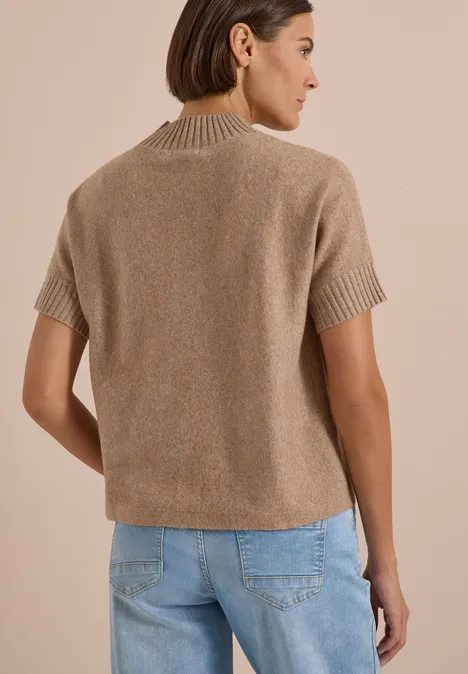 Frau von hinten in kurzem kastigem beige Strickpullover mit kurzen Ärmeln und Jeans vor beigem Hintergrund.