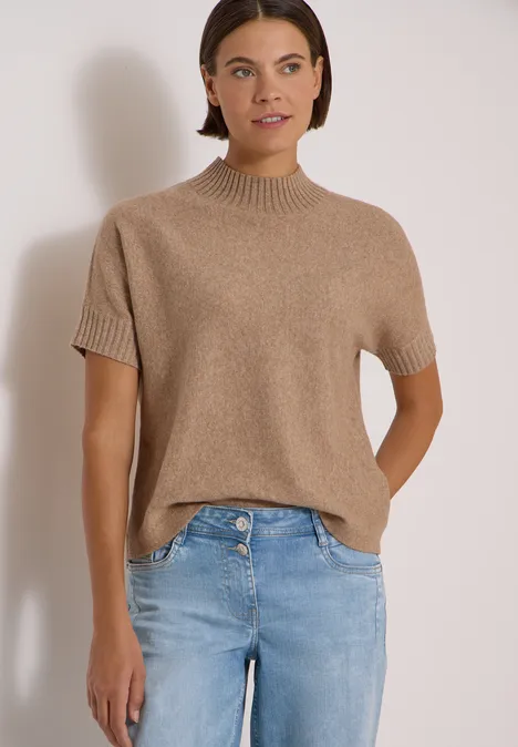Eine Frau trägt einen kurzärmligen beigen Strickpullover und blaue Jeans vor hellem Hintergrund.