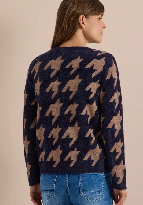 Frau von hinten in blau-beigem Strickpullover mit großem Hahnentritt-Muster und blauer Jeans vor hellem Hintergrund.