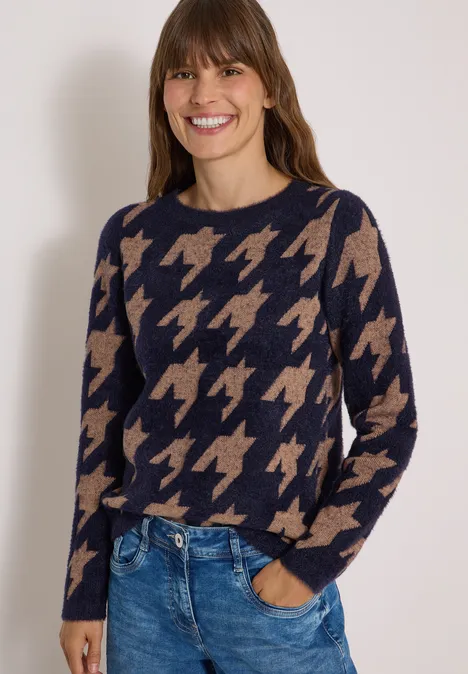 Frau trägt dunkelblauen Pullover mit beigem Hahnentrittmuster und Jeans, sie lächelt