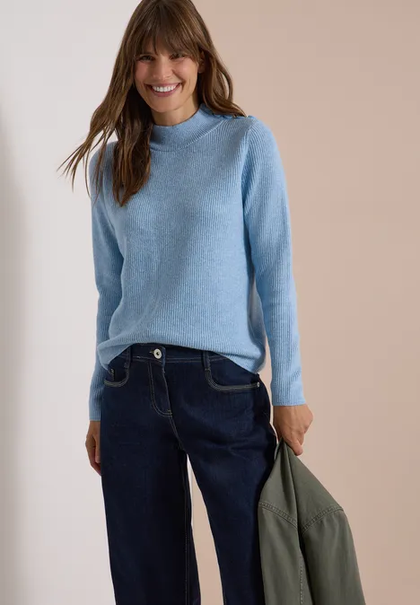 Pullover mit Struktur und Knopfdetail easy blue melange Frau in hellblauem Pullover und dunklen Jeans, hält grüne Jacke, lächelt vor neutralem Hintergrund.