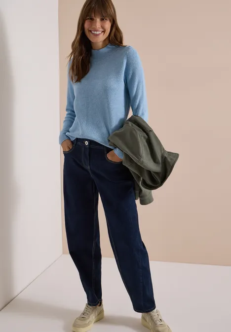 Pullover mit Struktur und Knopfdetail easy blue melange Junge Frau in hellblauem Pullover, dunklen Jeans und weißen Sneakern, hält grüne Jacke, lächelt.