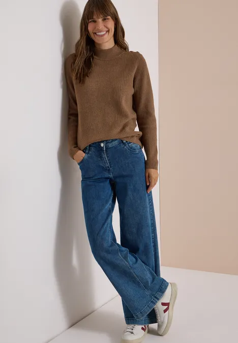 Pullover mit Struktur und Knopfdetail dark toffee brown melange Eine junge Frau lehnt lässig an einer Wand in braunem Pullover, blauen Jeans und Sneakers.