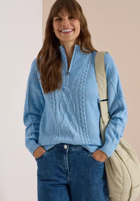 Lächelnde Frau in hellblauem Zopfpullover mit Reißverschluss, Jeans und beiger Tasche vor hellem Hintergrund.