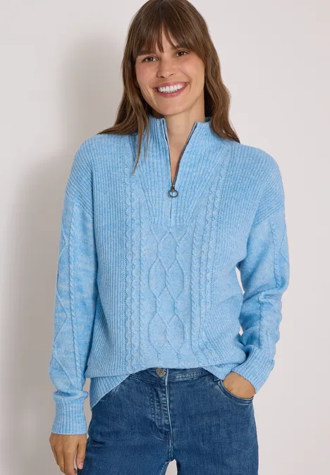 Frau mit blauem Strickpullover und Jeans, sie lächelt und hat die Hände in den Taschen.