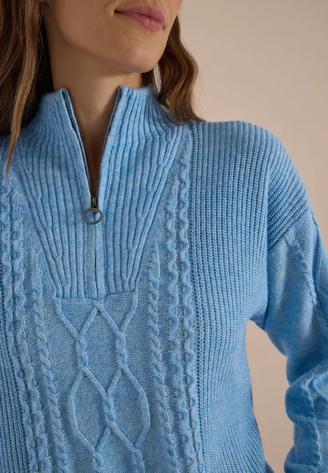 Frau trägt hellblauen Strickpullover mit Zopfmuster und halbem Reißverschluss am Kragen.
