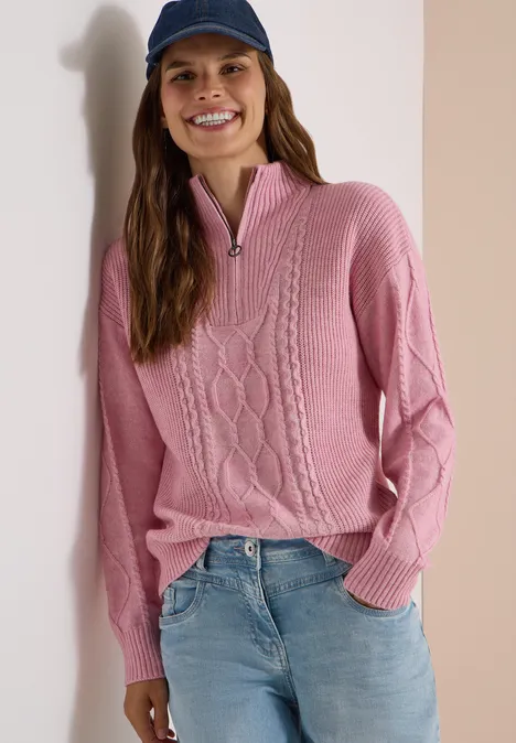 Eine lächelnde Frau in rosa Zopfmuster-Pullover mit Reißverschluss, Jeans und dunkelblauer Mütze.