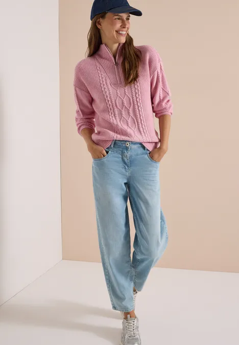 Frau in pinkem Strickpullover, blauer Jeanshose und Sneakern vor neutralem Hintergrund.