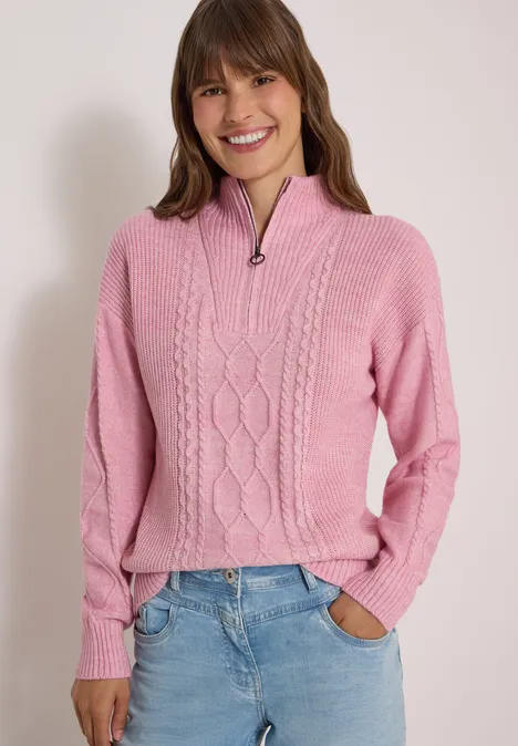 Eine lächelnde Frau in rosa Zopfmuster-Pullover mit halbem Reißverschluss und Jeans.