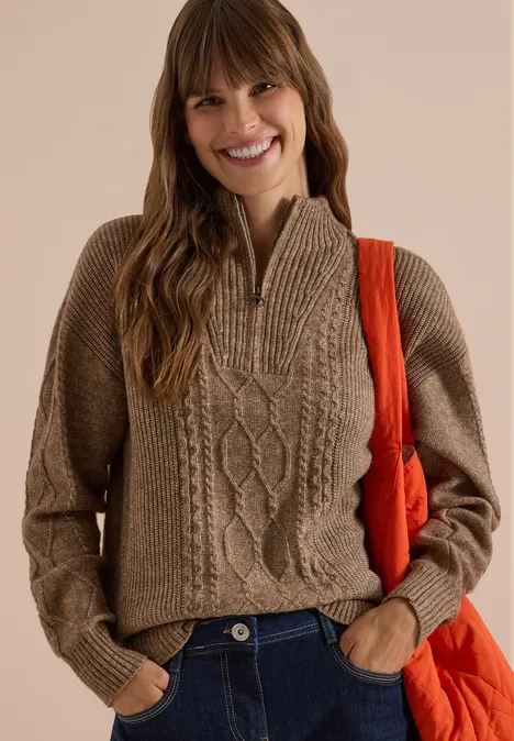 Eine Frau im braunen Zopfmuster-Strickpullover mit halbem Reißverschluss und orangefarbener Tasche, lächelnd.